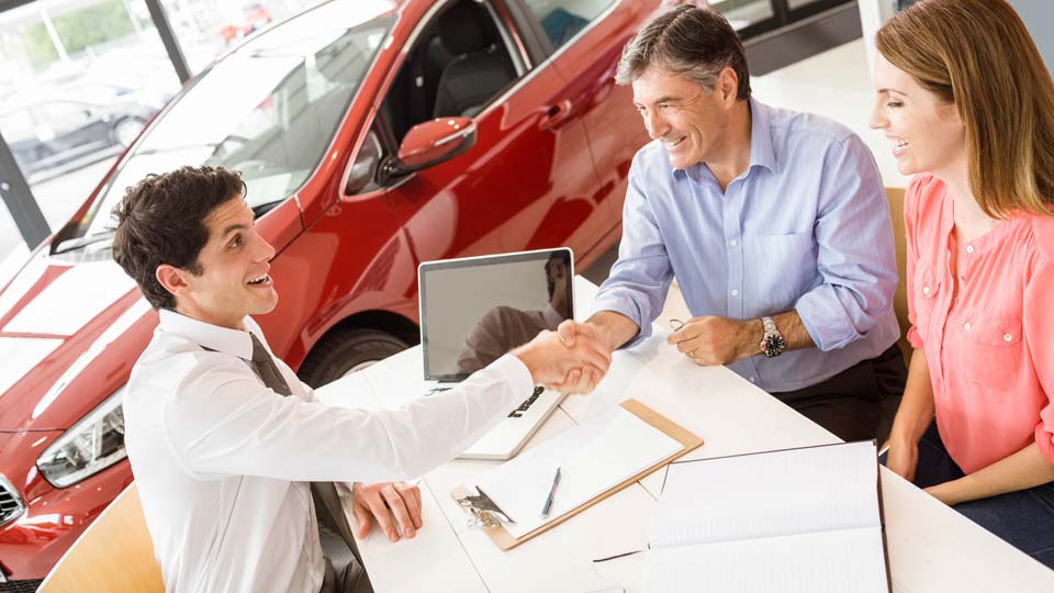 Ein Autoverkäufer schüttelt den Kunden die Hand in einem Autohaus in Braunschweig, im Hintergrund ist ein rotes Auto zu sehen und auf dem Tisch liegen Unterlagen.